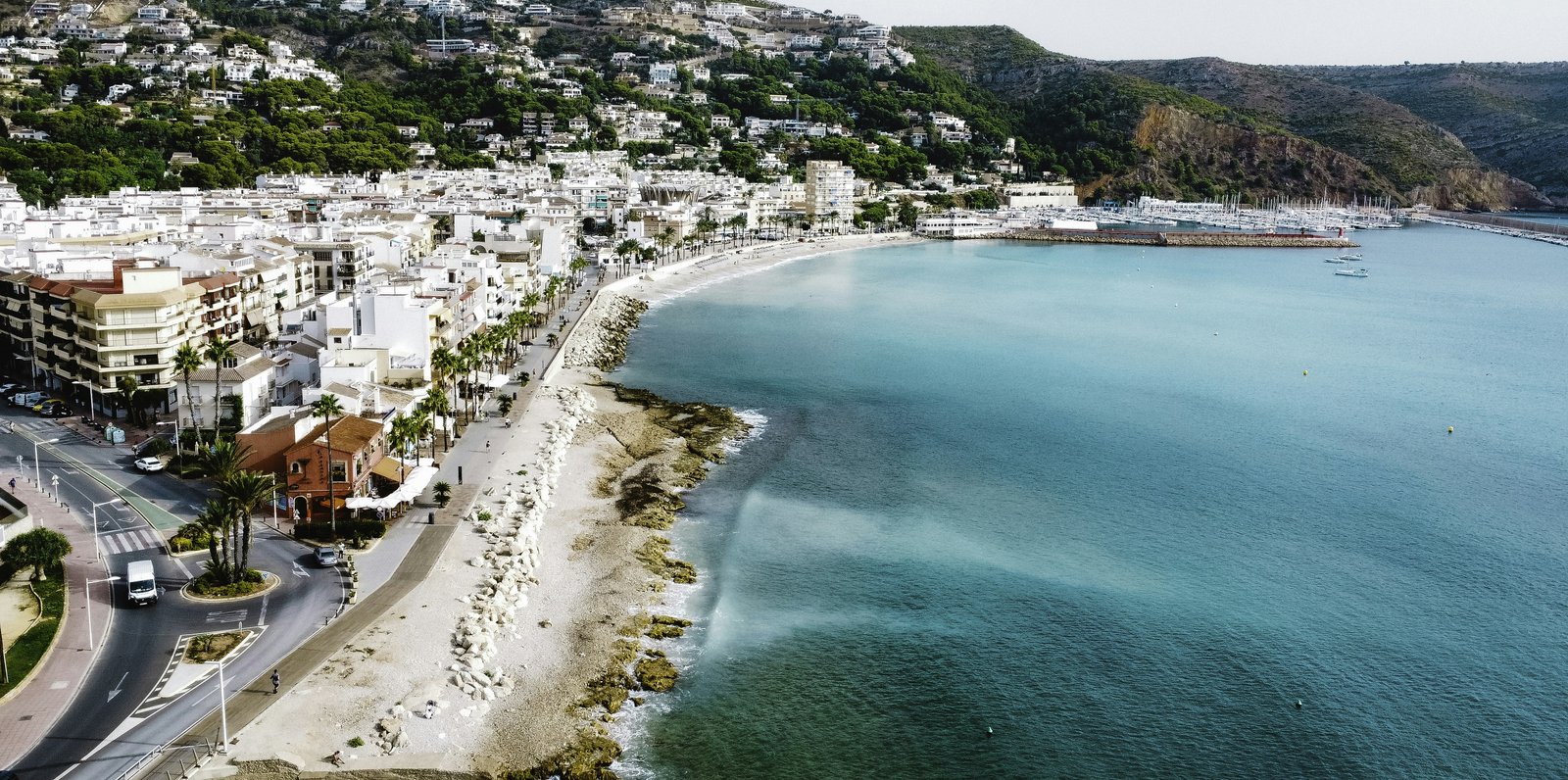 Luftaufnahme des Hafens von Jávea mit dem Kiesstrand La Grava und dem Yachthafen, dahinter die grüne Flanke des Cap de Sant Antoni mit den Villen von La Corona.